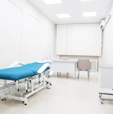 Bright, clean medical examination room with a teal examination table, white walls, desk, chair, and medical equipment, conveying a sterile, professional atmosphere.