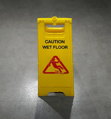 Yellow caution wet floor sign on a shiny gray surface, displaying a red pictogram of a person slipping. The tone is alert and safety-focused.