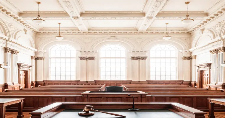 Elegant courtroom with tall arched windows, wooden decor, and ornate ceiling. A gavel on a desk conveys authority and formality. Bright and well-lit.