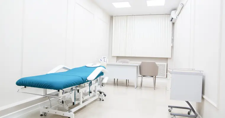 Bright, clean medical examination room with a teal examination table, white walls, desk, chair, and medical equipment, conveying a sterile, professional atmosphere.