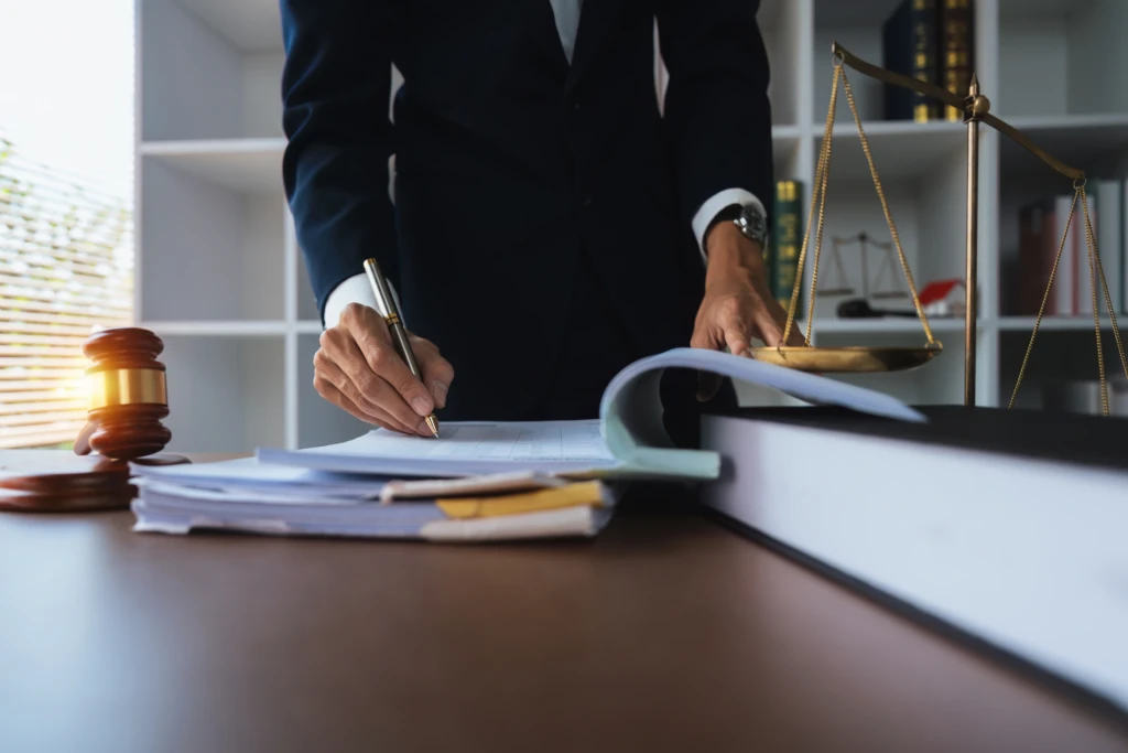 A person in a dark suit is writing on documents at a desk. Nearby are a gavel and scales of justice, conveying a legal setting and serious tone.
