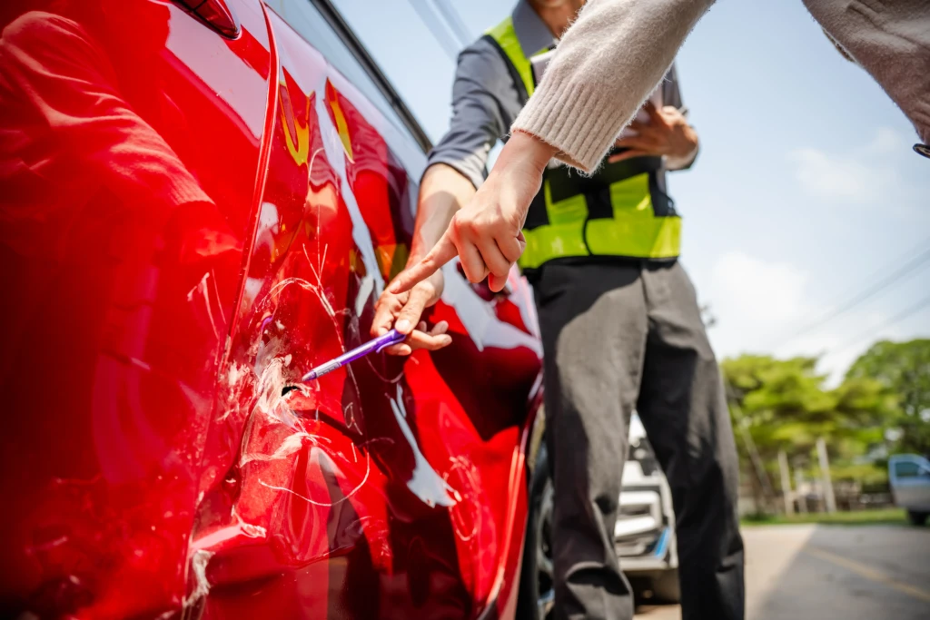 A close-up of a red car with a dent and scratches on the side. Two people, one in a reflective vest, examine the damage with a pen. Trees are in the background.