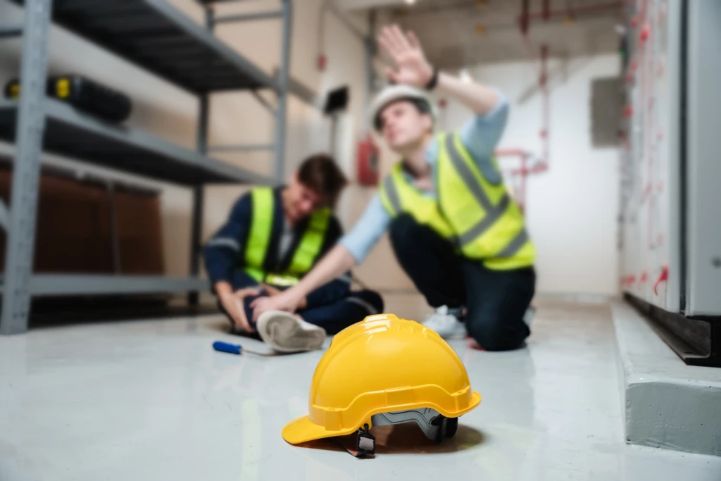 A yellow hard hat lies on the floor of an industrial setting. In the background, two workers in safety vests attend to an injured colleague.