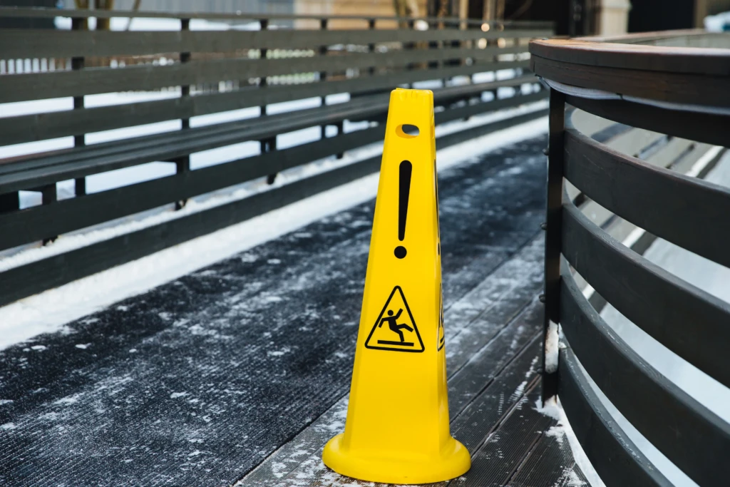 Yellow caution cone on an icy walkway with a slipping figure and exclamation mark. Black railings line the path, signaling a safety hazard.
